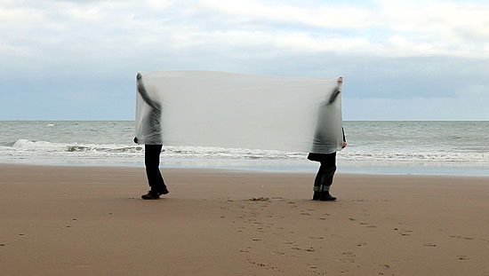 Two figures on a beach stretch a length of parachute silk between them, partly obscuring both their own bodies and the distant horizon