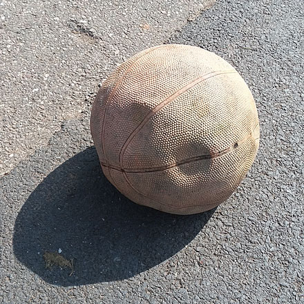 A slightly deflated basketball sits on tarmac in strong sunlight.