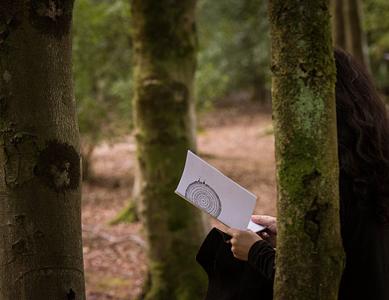 A dark-haired woman, dressed in dark clothing, stands among trees, reading from a white booklet. On its cover, the booklet has a diagram of a section through a tree trunk.