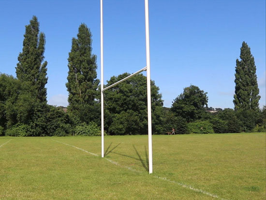 A view across the goal area of a rugby field, with tall trees and blue sky behind.  The posts are in strong sunlight and cast uncanny double shadows.