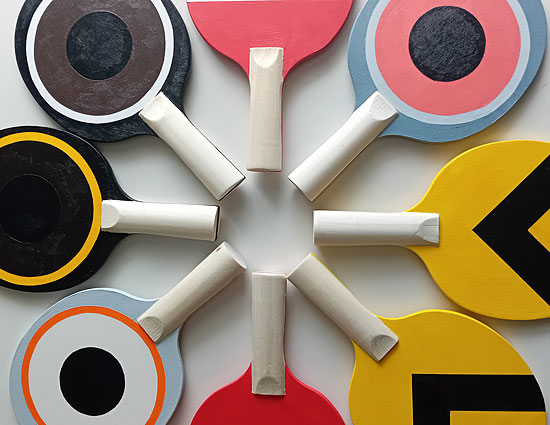 A face-forward image of eight table tennis bats arranged in a circle with their wooden handles meeting in the centre. Each face is painted with either a colourful railway signal or a birds eye